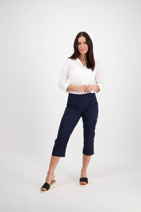 A woman with long dark hair stands against a white background, smiling softly. She wears a white button-up shirt, black slip-on sandals, and Vassalli Wave Pant in navy with an adjustable waistband. Her hands are lightly clasped.