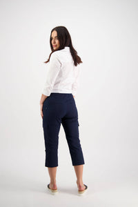A woman with long brown hair, facing backward and looking over her shoulder, wears a white shirt, black slip-on shoes, and Vassalli's Wave Pant in Navy against a plain white background.