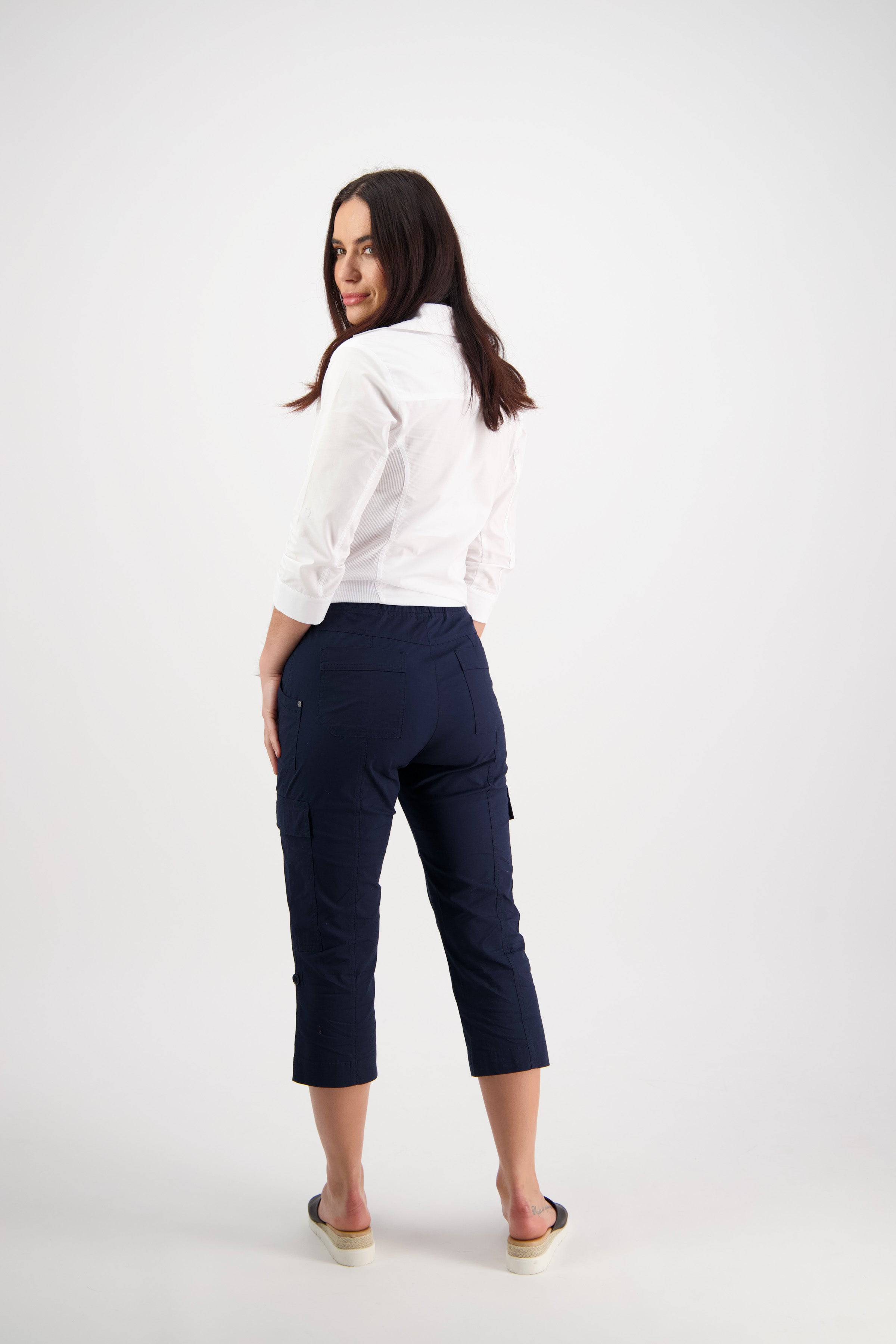 A woman with long brown hair, facing backward and looking over her shoulder, wears a white shirt, black slip-on shoes, and Vassalli's Wave Pant in Navy against a plain white background.
