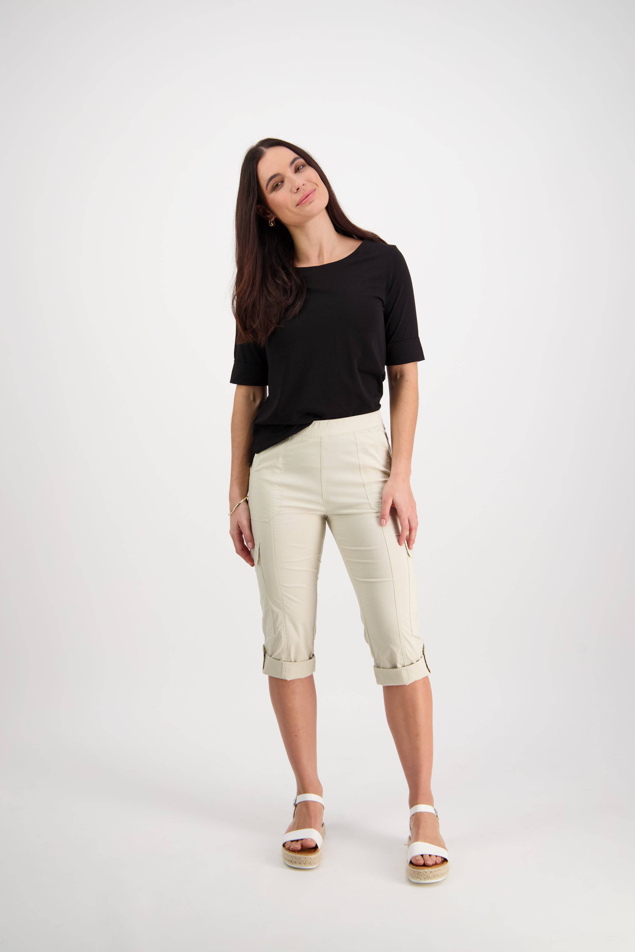 A woman with long brown hair smiles against a white background. She wears a black top, Vassalli Wave Pant - Sand with a pull-on waistband, and white sandals.