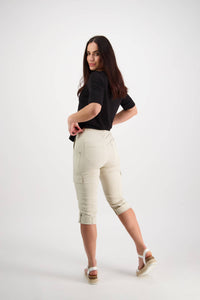 A woman with long dark hair stands against a white background, looking over her shoulder. She wears a black top, Vassalli Wave Pant - Sand, and white sandals, with one foot slightly raised behind her.