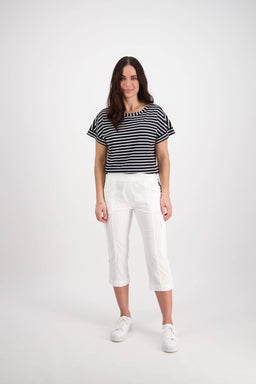 A woman with long brown hair smiles at the camera against a white background, wearing Vassalli’s Wave Pant in white, a black and white striped T-shirt, and white sneakers.