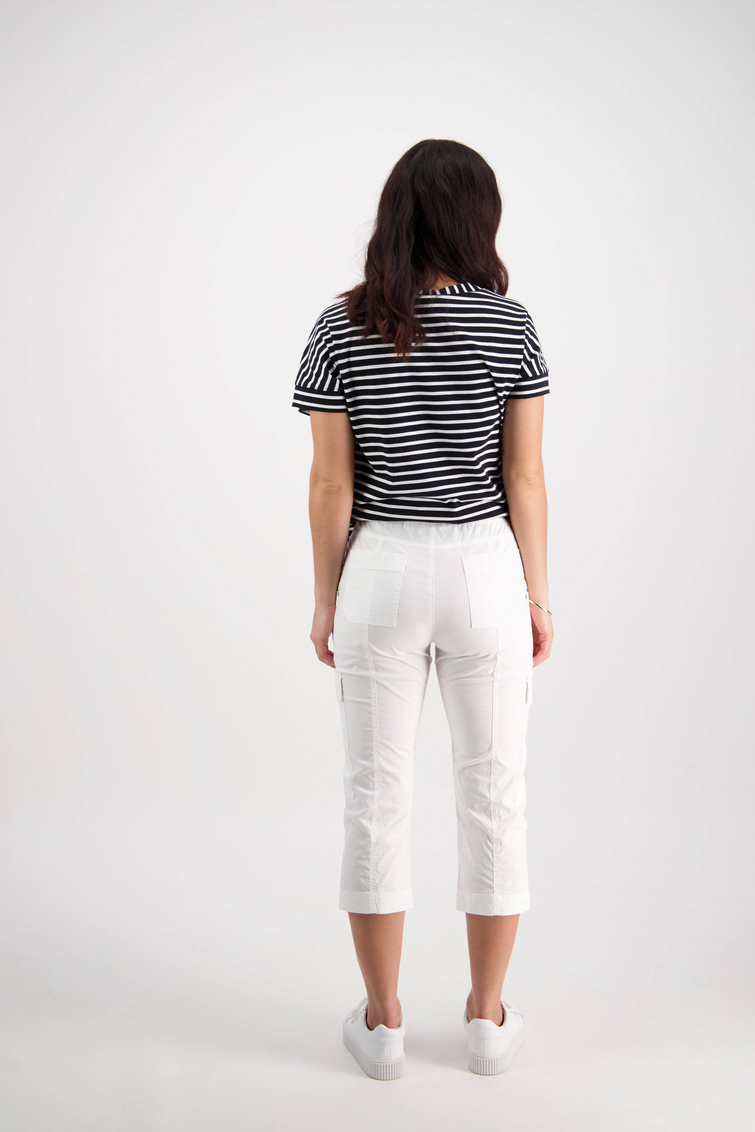 A woman with long dark hair wears a black and white striped short-sleeve shirt, Vassalli Wave Pant - White, and white shoes, standing facing away from the camera against a plain light background.