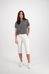 A woman with long brown hair stands in a studio wearing Vassalli's Wave Pant - White and a black and white striped t-shirt, paired with white sneakers. She smiles slightly with one hand in her pocket against a plain white background.