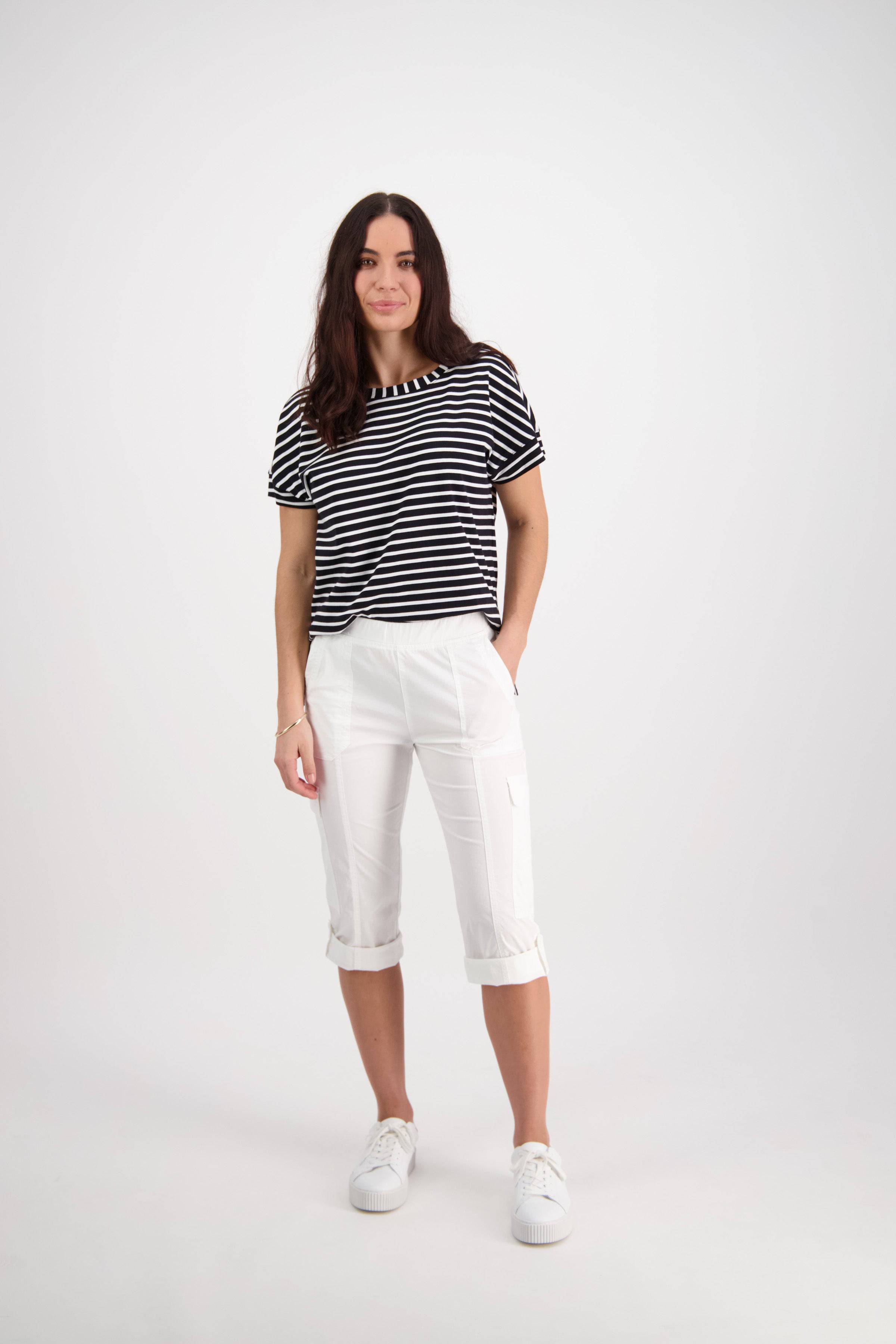 A woman with long brown hair stands in a studio wearing Vassalli's Wave Pant - White and a black and white striped t-shirt, paired with white sneakers. She smiles slightly with one hand in her pocket against a plain white background.