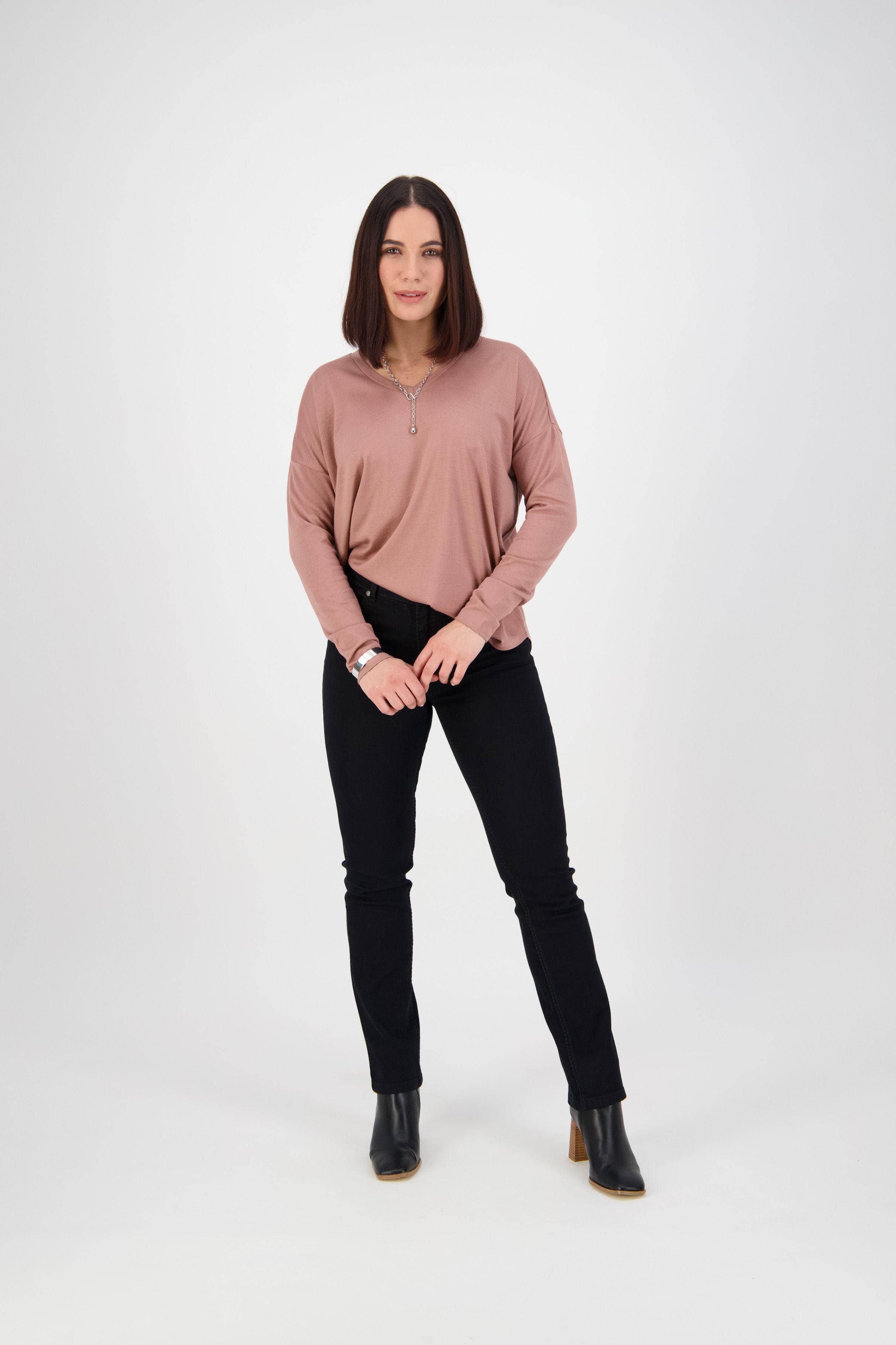 A woman with straight brown hair faces the camera, wearing a mauve long-sleeve top, Vassalli Lotus Jean in black denim, and black ankle boots. She stands with her hands clasped in front of her against a plain white background.