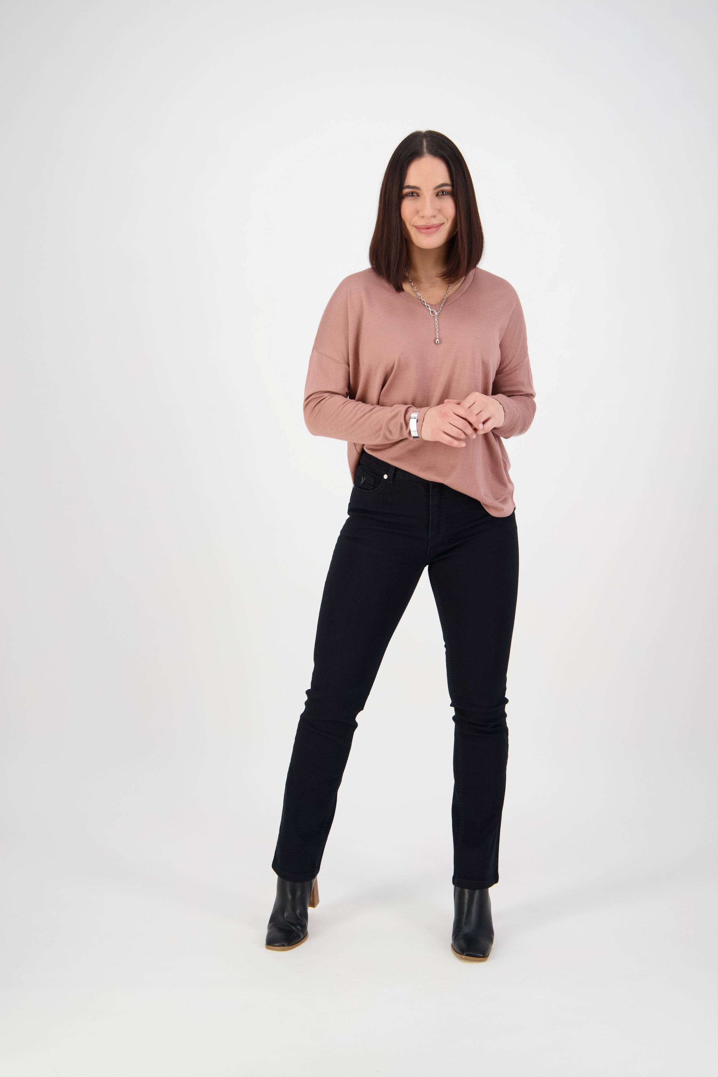 A woman with straight, shoulder-length dark hair stands against a white background, wearing a pink long-sleeve top, Vassalli Lotus Jean - Black Denim, and black ankle boots. She smiles softly with relaxed hands.