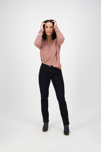 A woman stands against a white background wearing a pink long-sleeve shirt, Vassalli Lotus Jean in black denim, and black ankle boots, with her hands touching her head and a slight smile.