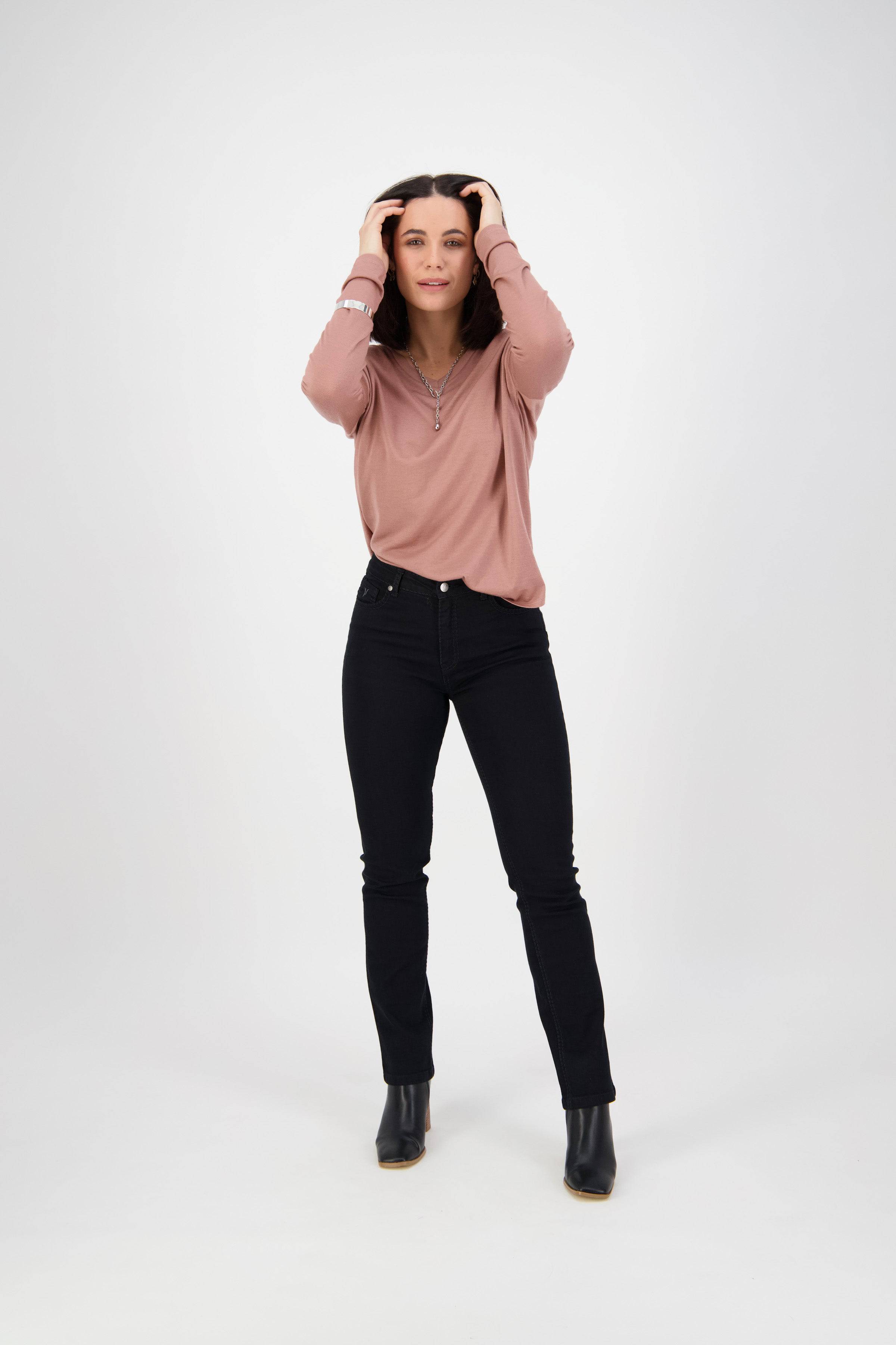 A woman stands against a white background wearing a pink long-sleeve shirt, Vassalli Lotus Jean in black denim, and black ankle boots, with her hands touching her head and a slight smile.