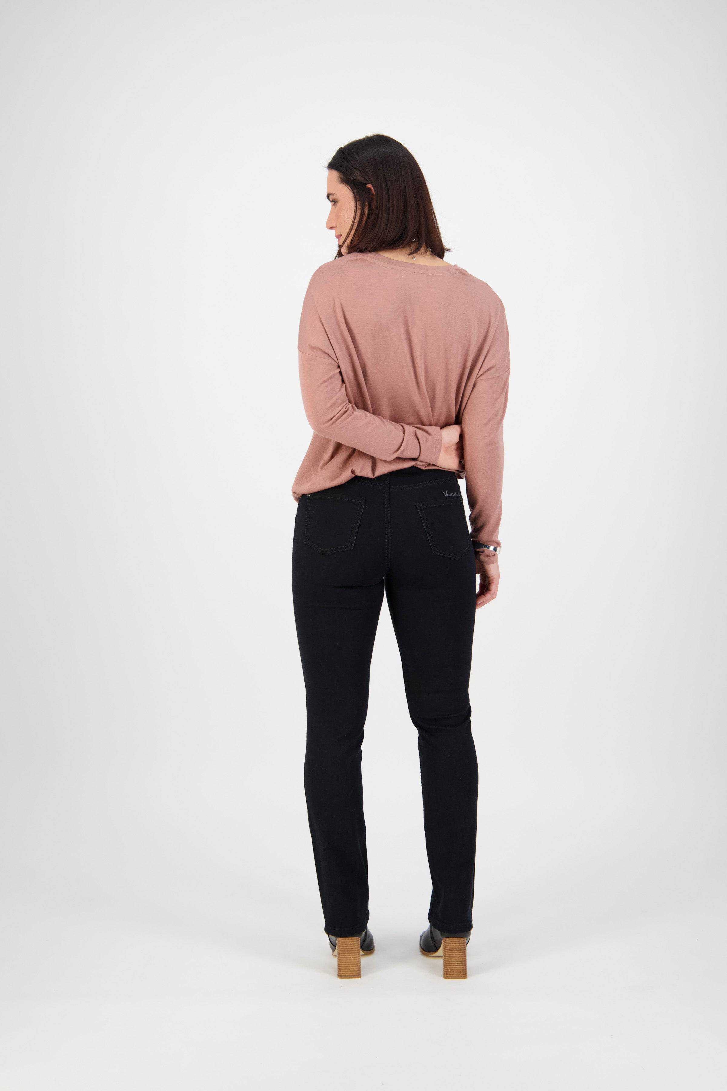 A woman with shoulder-length brown hair, seen from behind in a minimalist studio, wears a mauve long-sleeve top, Vassalli Lotus Jean - Black Denim, and tan heeled shoes with her right arm behind her back.