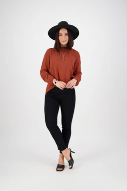 A woman in a wide-brimmed black hat, rust-colored long-sleeve top, and Vassalli Icon Jean in Black Denim stands against a plain white background, looking at the camera with a slight smile.