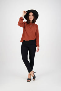 A woman stands against a white background, slightly tipping her hat, wearing a black wide-brim hat, rust long-sleeve top, Vassalli Icon Jean in black denim, and black heeled sandals.