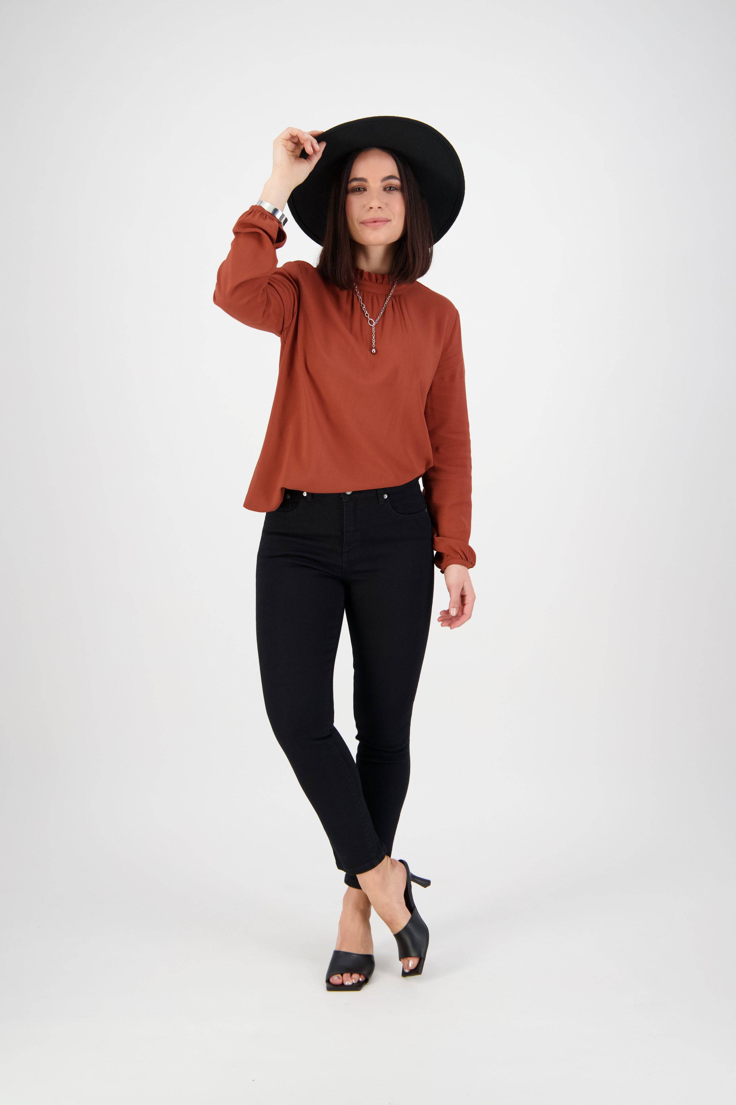 A woman stands against a white background, slightly tipping her hat, wearing a black wide-brim hat, rust long-sleeve top, Vassalli Icon Jean in black denim, and black heeled sandals.