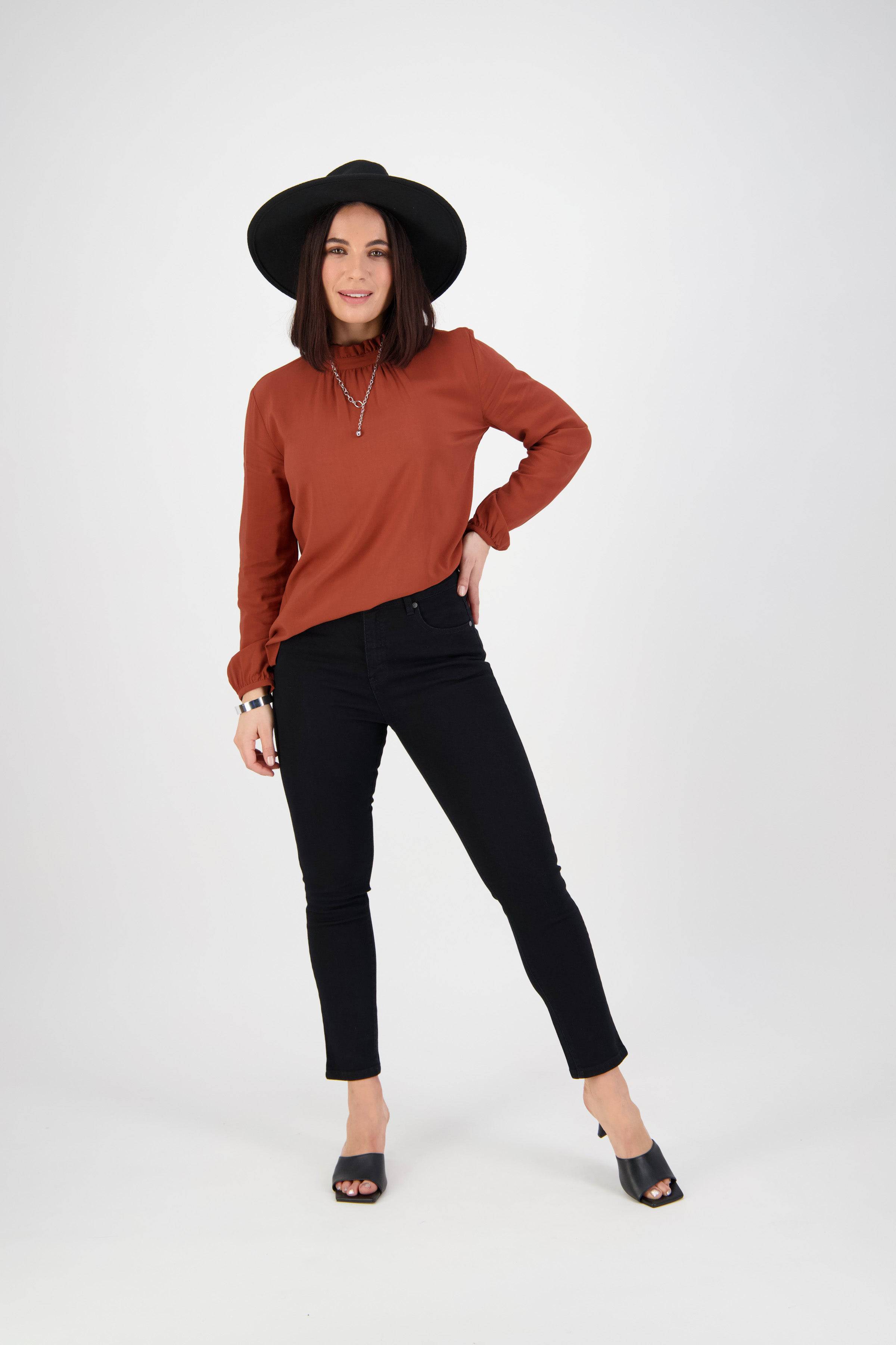 A woman stands against a white background in a black wide-brim hat, rust long-sleeve blouse, Vassalli Icon Jean - Black Denim skinny jeans, and black heeled sandals. She smiles and poses with one hand on her hip.