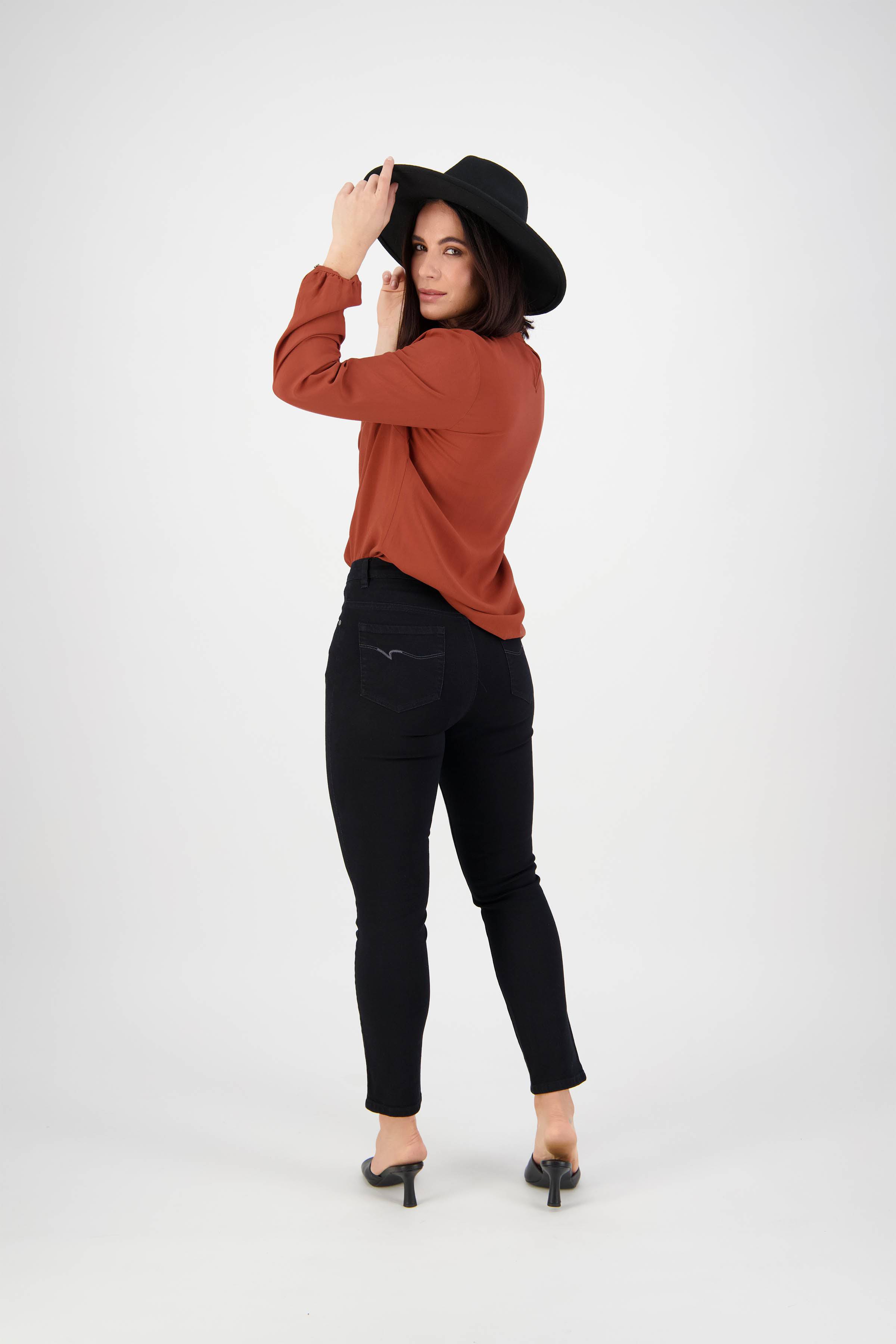A woman in a black hat, rust long-sleeve blouse, and Vassalli Icon Jean - Black Denim stands with her back to the camera on a plain white background, looking over her shoulder and touching the brim of her hat.