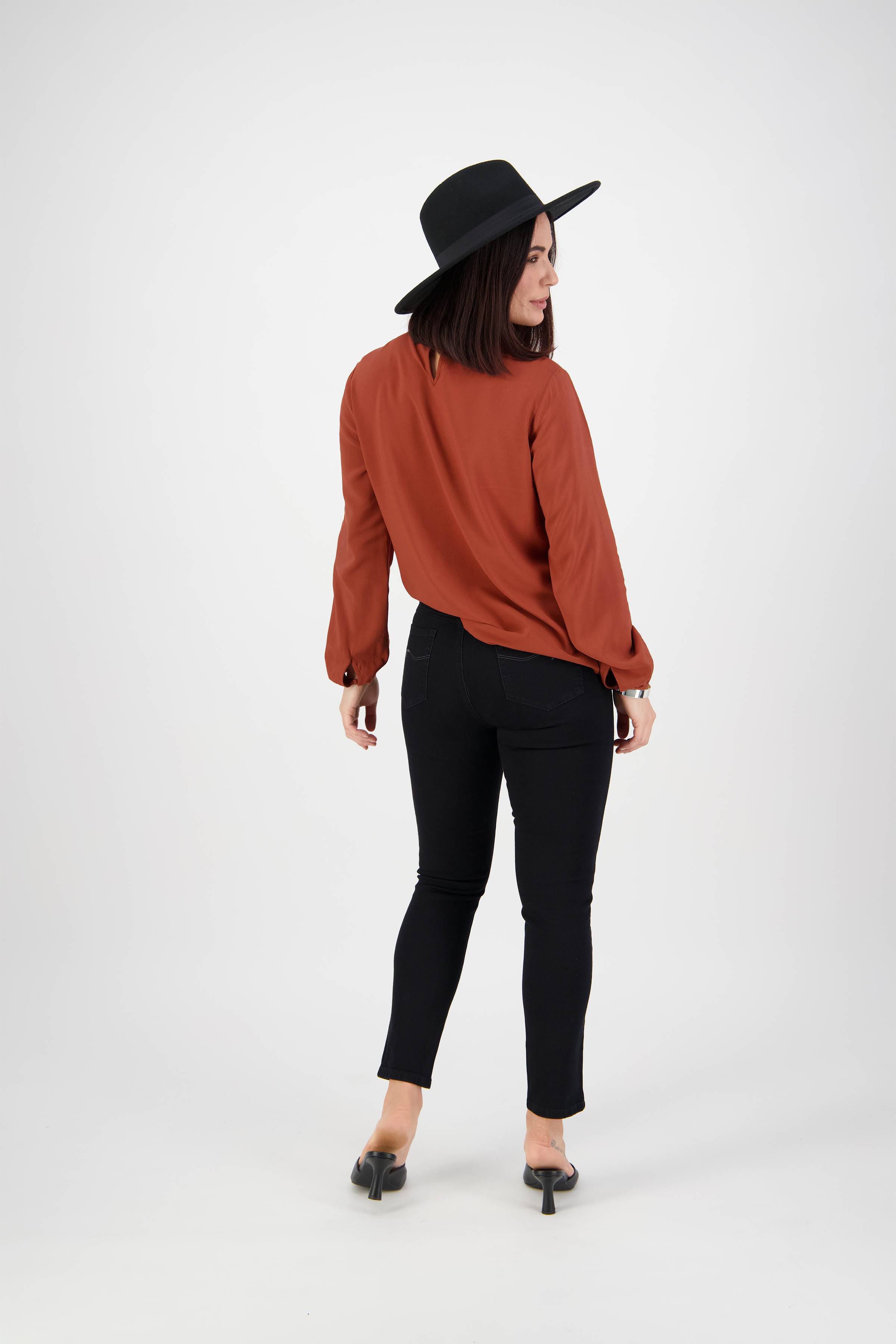 A woman stands facing away against a plain white background, wearing a black wide-brimmed hat, a rust long-sleeve blouse, and the Vassalli Icon Jean in Black Denim.