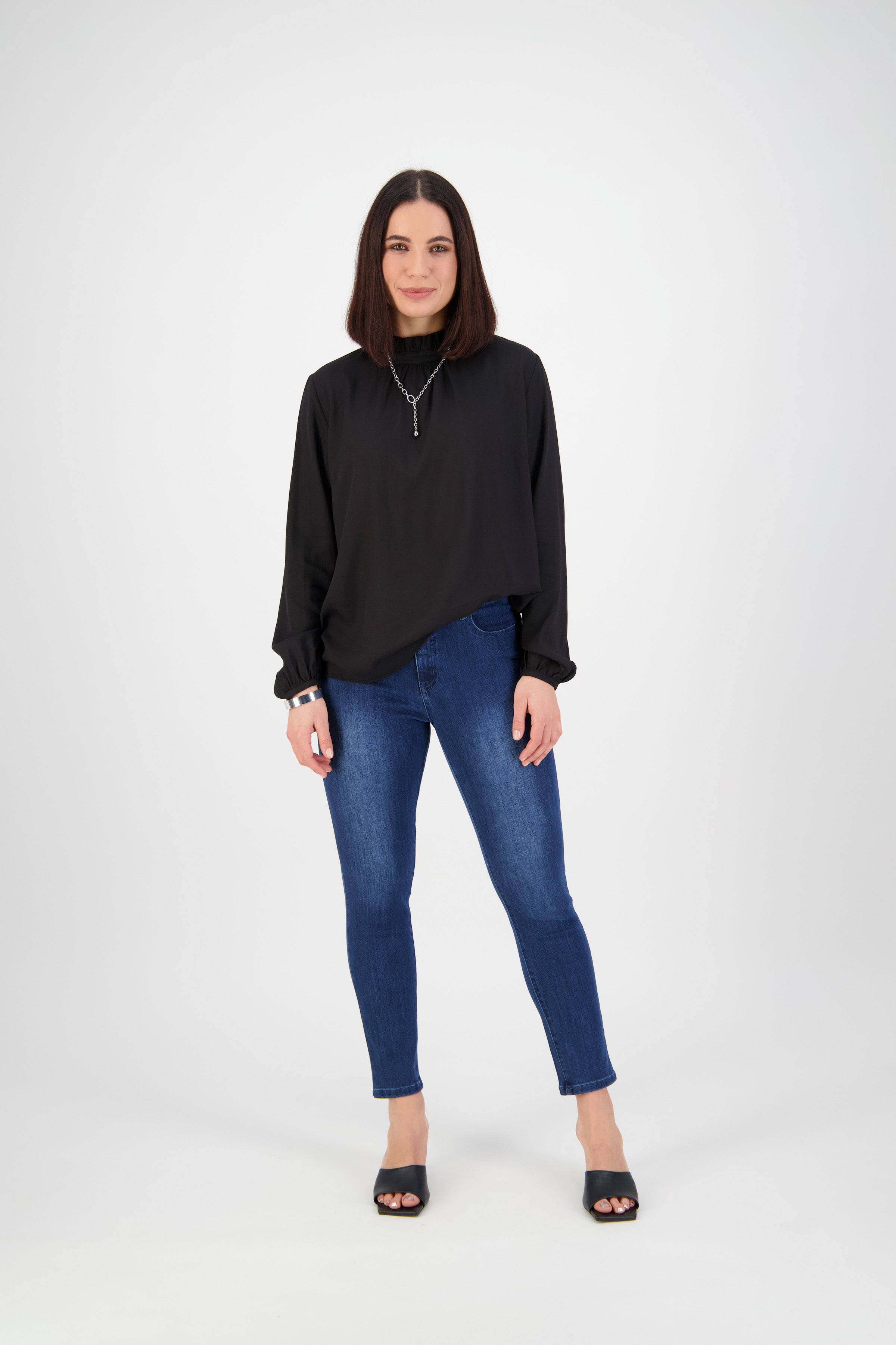 A woman with straight, shoulder-length dark hair stands against a plain white background, wearing Vassalli's Icon Jean in Blue Denim, paired with a black long-sleeve top, black open-toe heels, and a necklace. She looks at the camera with a slight smile.