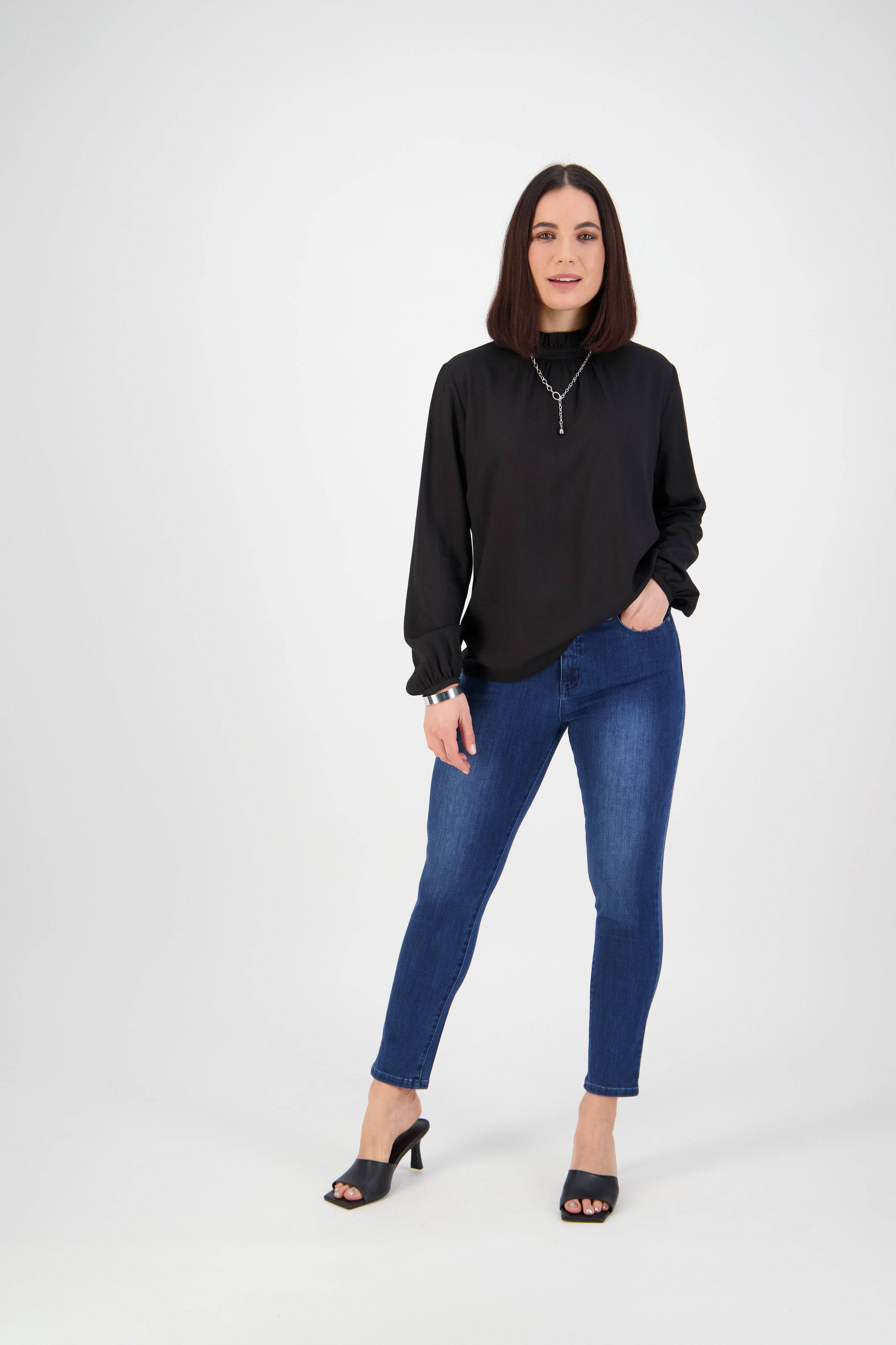 A woman with straight brown hair stands against a white background, wearing Vassalli’s Icon Jean - Blue Denim, a black long-sleeve blouse, black heeled sandals, and a necklace. One hand is in her pocket and she smiles slightly.