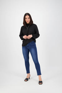 A woman with straight dark hair wears the Vassalli Icon Jean in Blue Denim, a black long-sleeve top, and black heeled sandals as she stands against a plain white background, looking at the camera and smiling slightly.