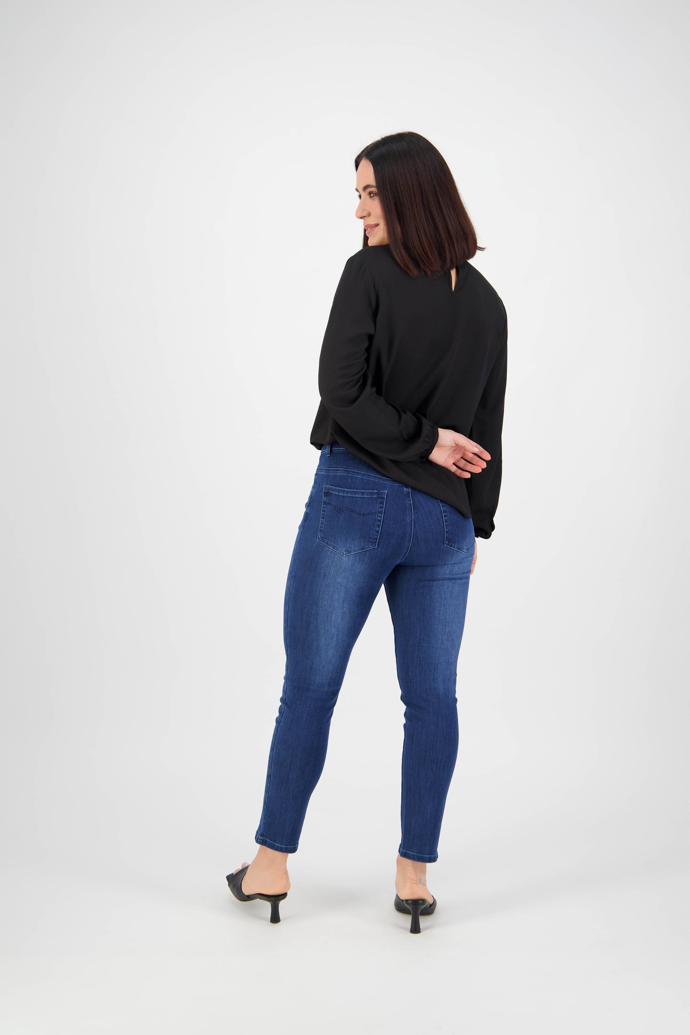 A woman with straight dark hair, dressed in a black long-sleeve top and Vassalli Icon Jean - Blue Denim, stands facing away and looks over her shoulder in a studio with a white background.