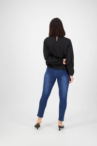 A woman faces away from the camera, wearing Vassalli Icon Jean - Blue Denim, a black long-sleeve top, and black heels against a plain white background.