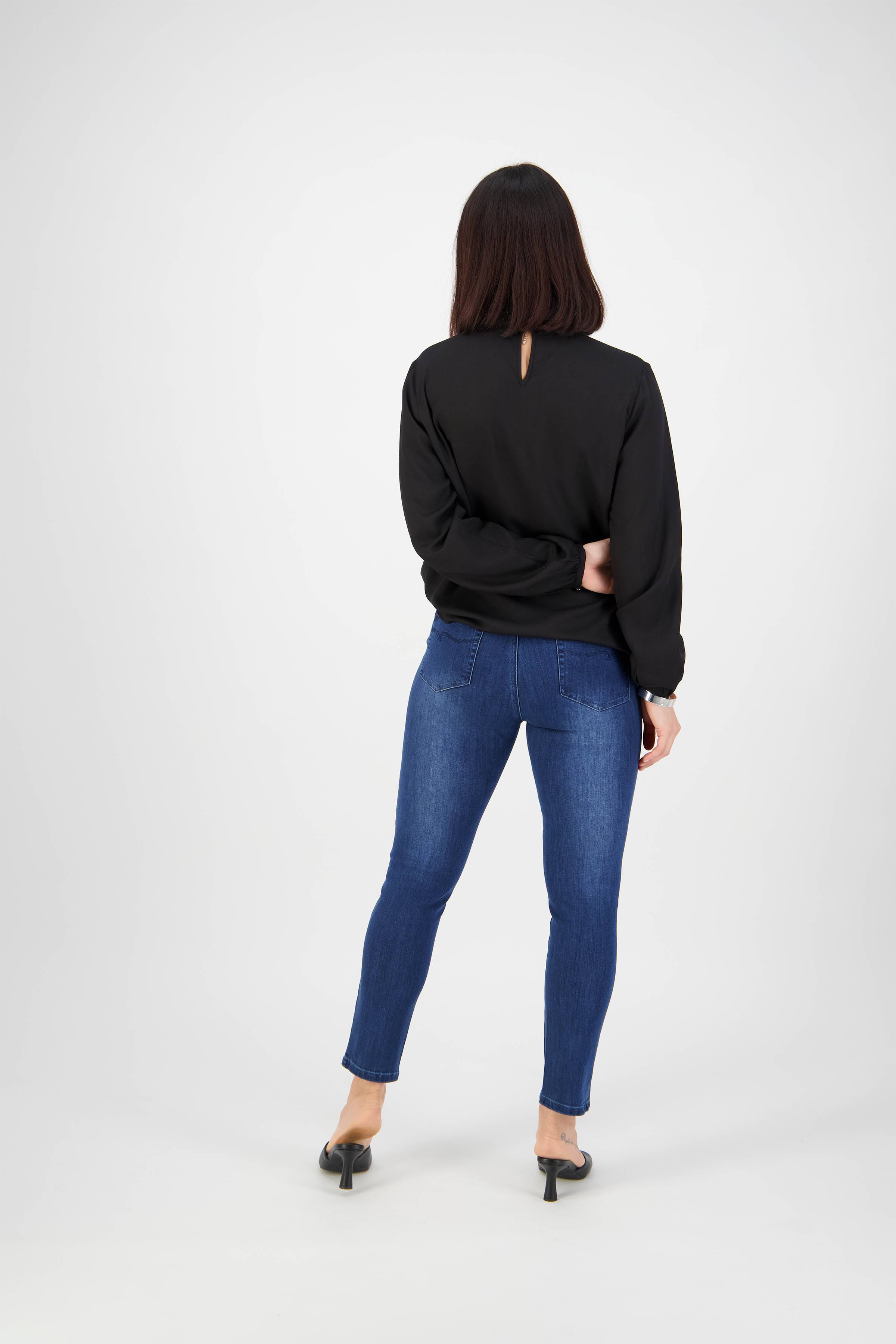 A woman faces away from the camera, wearing Vassalli Icon Jean - Blue Denim, a black long-sleeve top, and black heels against a plain white background.