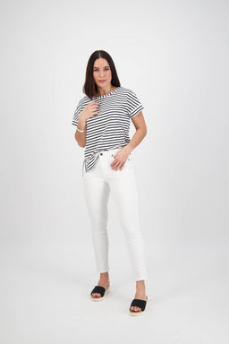 A woman stands in a studio wearing Vassalli’s Icon Jean in White Denim with a black and white striped t-shirt and black sandals. She has straight, shoulder-length hair, smiles slightly, and poses against a plain white background.