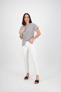 A woman poses against a plain white background wearing the Vassalli Icon Jean in White Denim, styled with a black and white striped t-shirt, black sandals, and a bracelet—one hand on her shirt, the other in her pocket.
