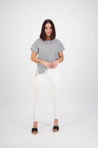 A woman with straight shoulder-length hair stands against a white background wearing Vassalli’s Icon Jean in White Denim, a black-and-white striped shirt, and black sandals. She looks at the camera with a slight smile.