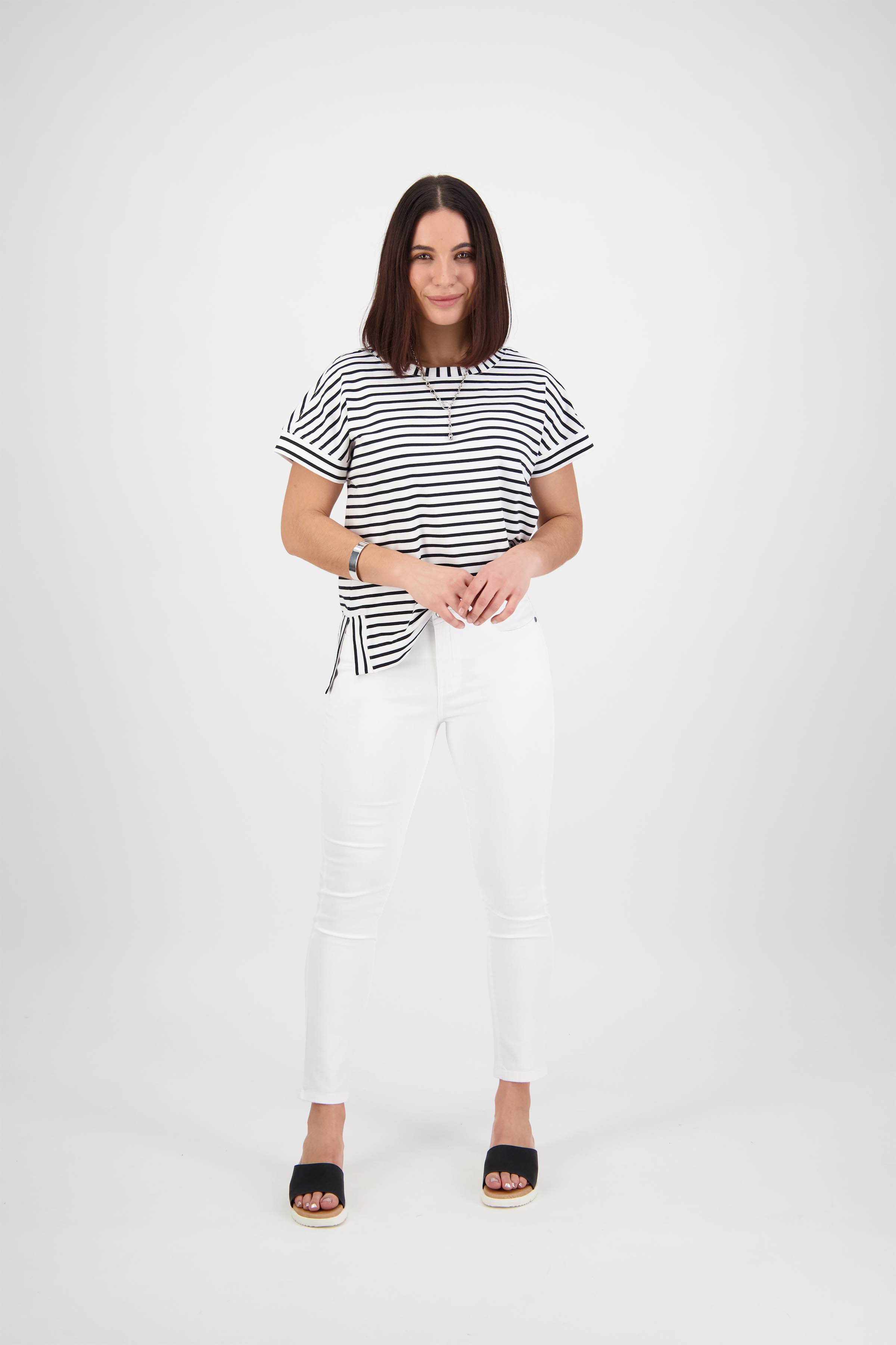 A woman with straight shoulder-length hair stands against a white background wearing Vassalli’s Icon Jean in White Denim, a black-and-white striped shirt, and black sandals. She looks at the camera with a slight smile.