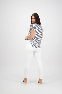 A woman with straight dark hair looks back over her shoulder, wearing Vassalli's Icon Jean in White Denim, a black-and-white striped tee, and black slip-on shoes against a plain white background.