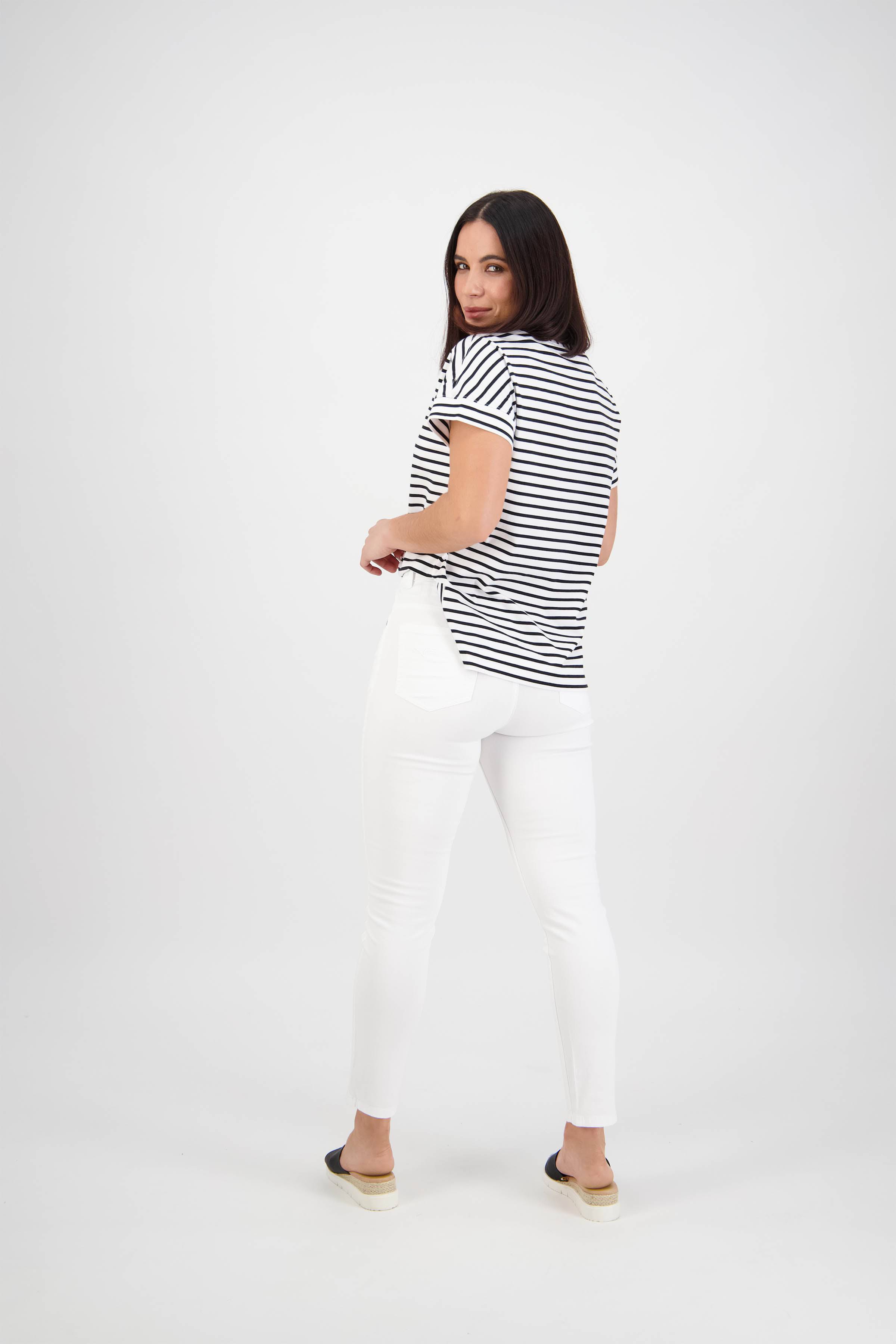 A woman with straight dark hair looks back over her shoulder, wearing Vassalli's Icon Jean in White Denim, a black-and-white striped tee, and black slip-on shoes against a plain white background.