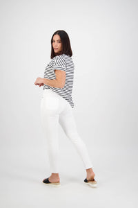 A woman with shoulder-length dark hair models the Vassalli Icon Jean in white denim, paired with a black and white striped t-shirt and black slip-on shoes, posing against a plain white background, looking over her shoulder.
