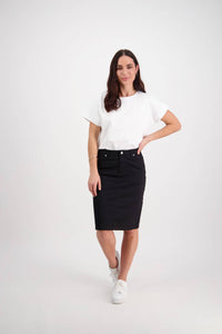 A woman stands against a white background, wearing the Vassalli Tilt Skirt in black, a white t-shirt, and white sneakers. She has long brown hair, one hand on her hip, and is smiling slightly.