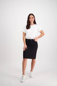 A woman with long dark hair and a neutral expression poses with one hand on her hip, wearing a white t-shirt, white sneakers, and the Vassalli Tilt Skirt in black against a plain white background.