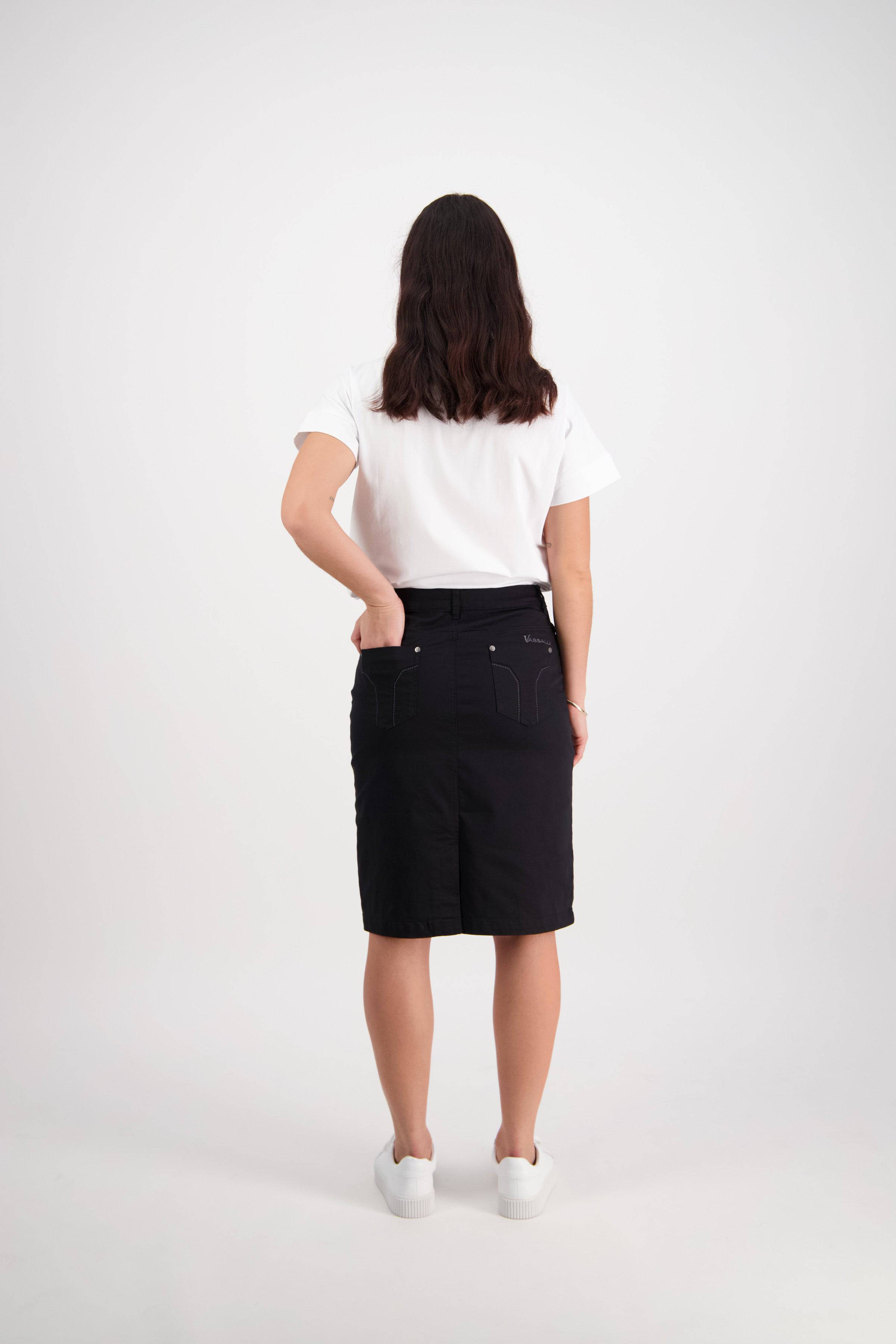 A person with long dark hair stands facing away against a light background, wearing the Vassalli Tilt Skirt in black, paired with a white T-shirt and white shoes, with one hand on their hip.
