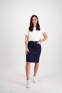 A woman with long brown hair, wearing a white t-shirt, white sneakers, and the Vassalli Tilt Skirt in Navy, stands against a plain white background with one hand in her skirt pocket.