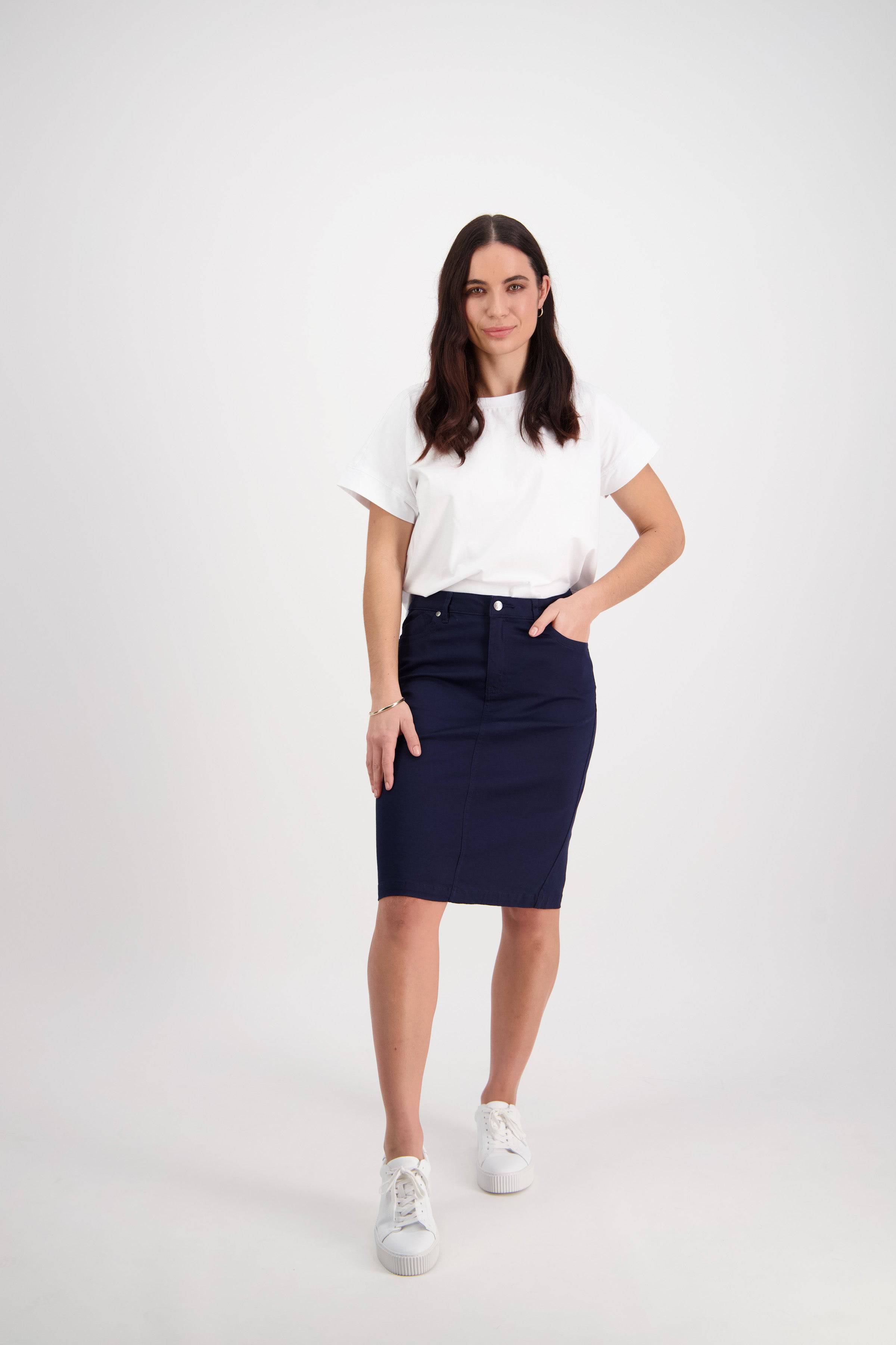 A woman with long brown hair, wearing a white t-shirt, white sneakers, and the Vassalli Tilt Skirt in Navy, stands against a plain white background with one hand in her skirt pocket.