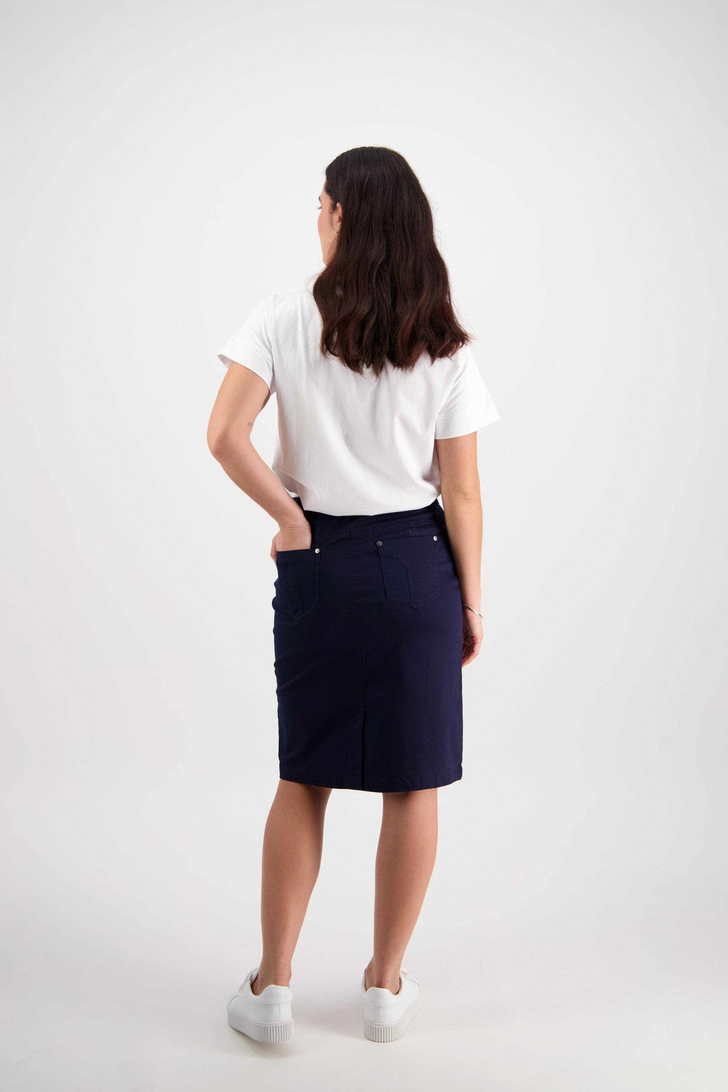 A woman with long dark hair faces away, wearing a white tee, Vassalli Tilt Skirt - Navy, and white sneakers against a plain light background. Her left hand rests on her hip and her right arm is relaxed. Ideal for everyday style.
