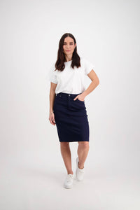 A woman with long dark hair stands against a white background, wearing a white t-shirt, Vassalli's Tilt Skirt in navy, and white sneakers. One hand is in her pocket as she looks at the camera.