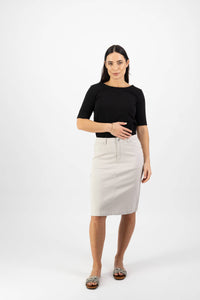 A woman with straight dark hair and a neutral expression wears a black short-sleeve top, Vassalli's Tilt Skirt in Pumice—a light beige, everyday skirt—and patterned flat sandals, standing against a plain white background.