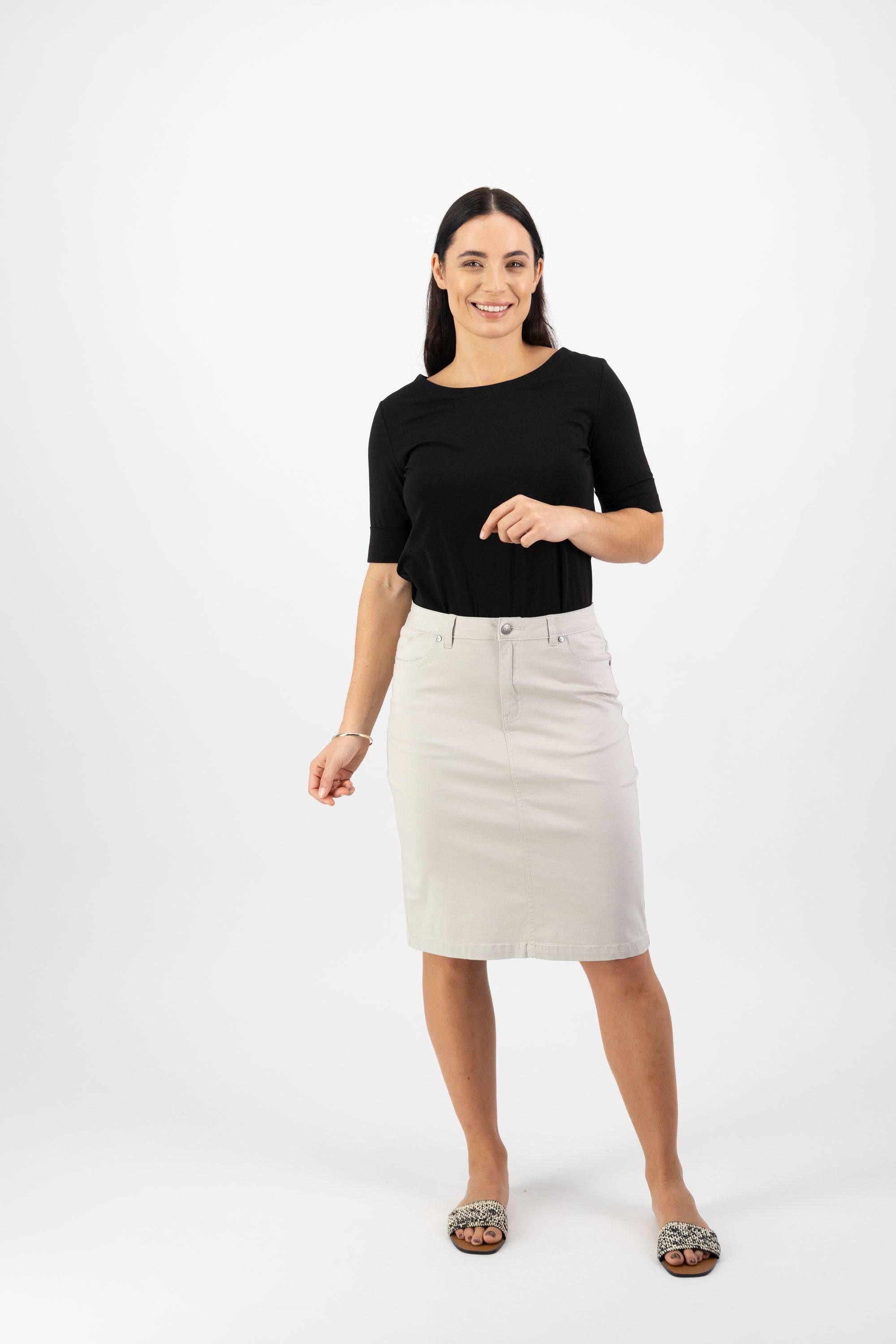 A woman with long dark hair, wearing a black top, Vassalli Tilt Skirt in Pumice, and patterned sandals, stands smiling against a plain white background.