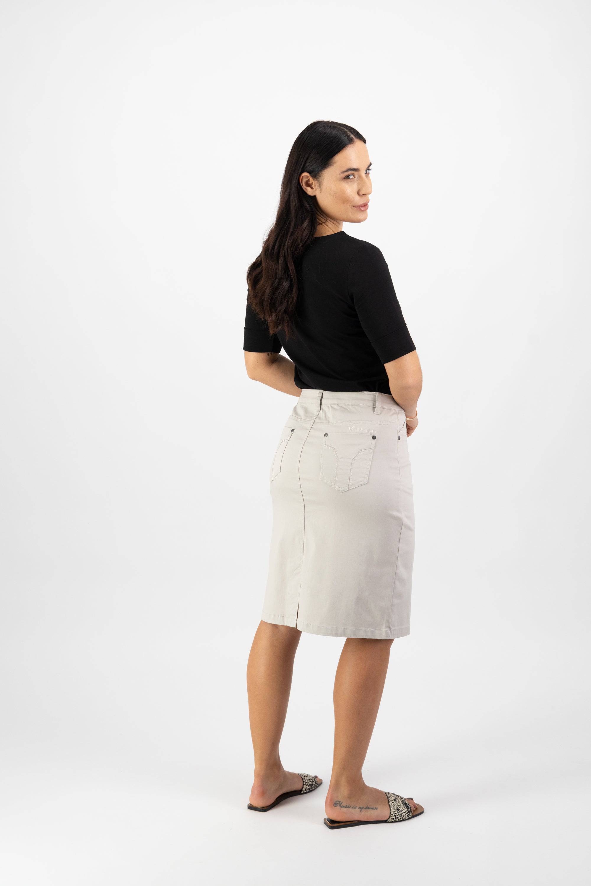 A woman with long dark hair, wearing a black top, Vassalli's Tilt Skirt in Pumice, and slide sandals stands facing away and looks back over her shoulder against a plain white background.