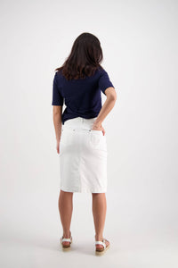 A woman faces away in a navy blue top and the Vassalli Tilt Skirt - White, paired with white sandals, one hand on her hip against a plain white background.