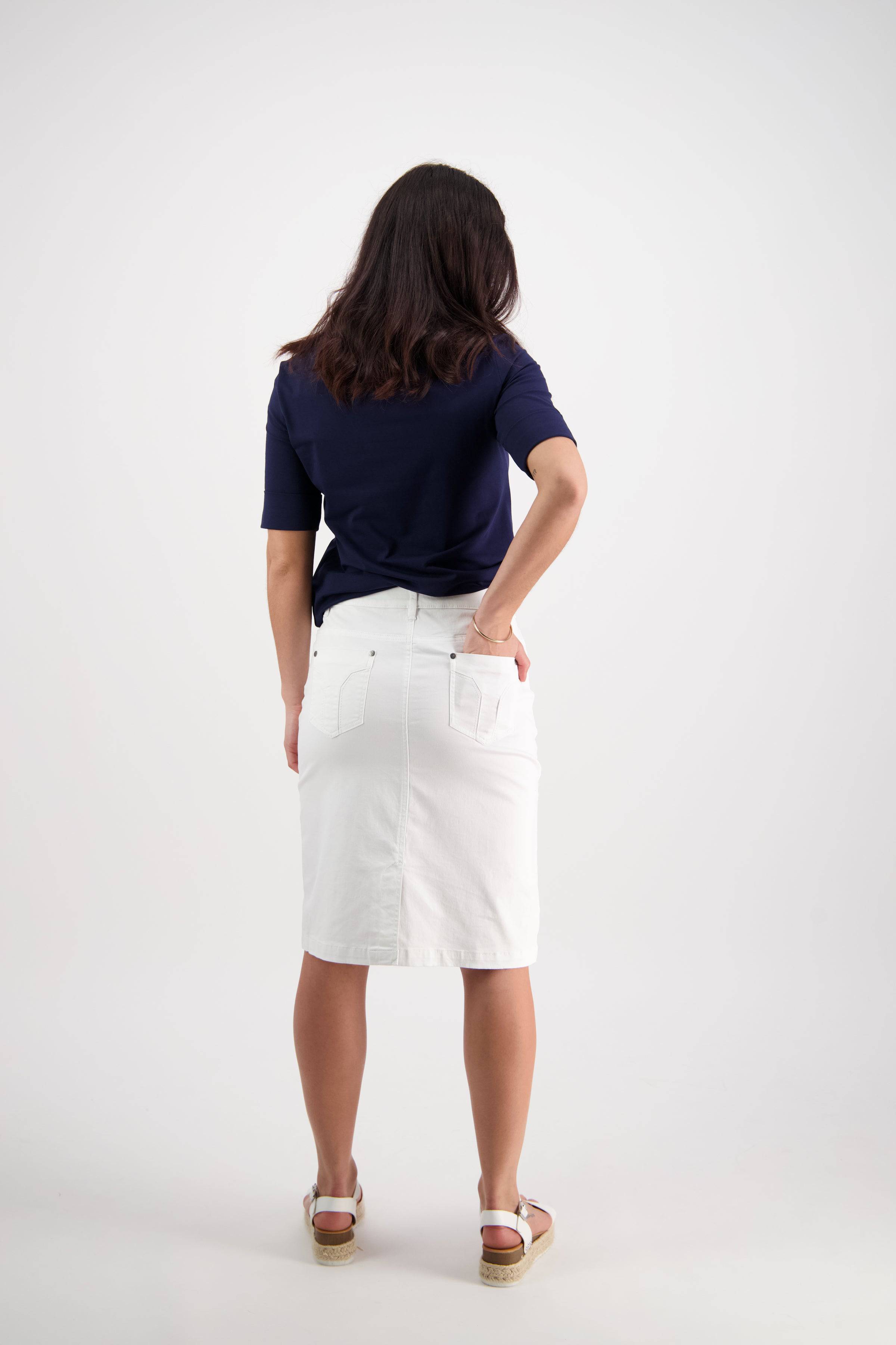 A woman faces away in a navy blue top and the Vassalli Tilt Skirt - White, paired with white sandals, one hand on her hip against a plain white background.