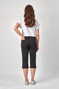 A woman with long wavy brown hair, wearing a white short-sleeve blouse, Vassalli 3/4 Pant - Black, and white sneakers, stands facing away from the camera against a plain light background.