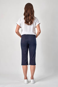 A woman with long wavy brown hair faces away, wearing a white short-sleeve shirt, Vassalli 3/4 Pant in navy, and white sneakers against a plain light gray background.