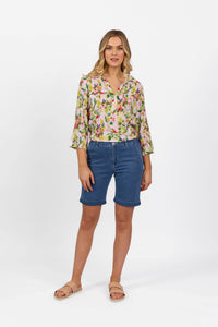 A woman with shoulder-length blonde hair and a relaxed expression wears Vassalli’s Havana Short in Blue Hue Denim, teamed with a floral three-quarter sleeve blouse and tan slide sandals, standing against a white background.