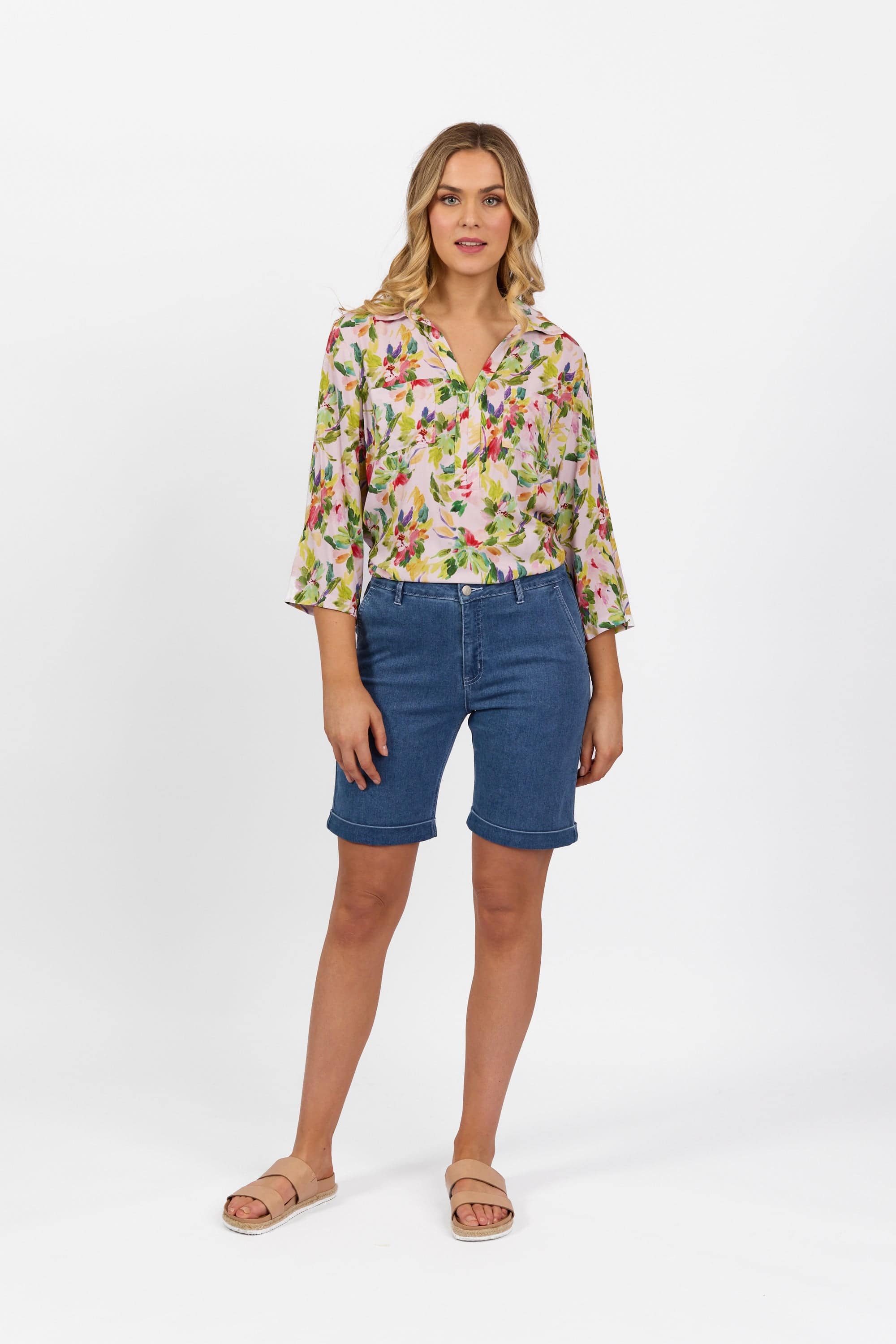 A woman with shoulder-length blonde hair and a relaxed expression wears Vassalli’s Havana Short in Blue Hue Denim, teamed with a floral three-quarter sleeve blouse and tan slide sandals, standing against a white background.