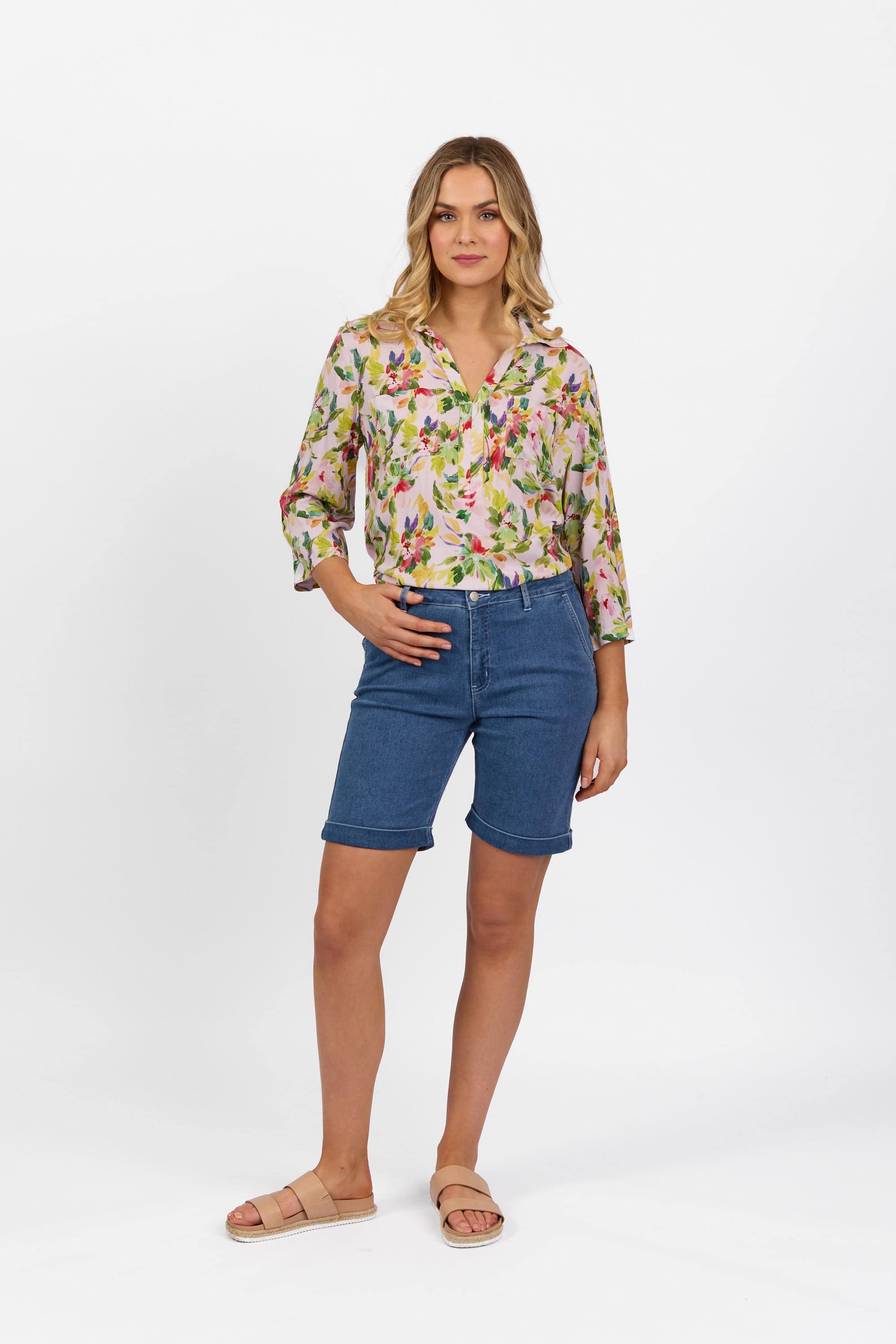 A woman stands against a white background wearing blue Havana Short - Blue Hue Denim by Vassalli, a floral button-up shirt with rolled sleeves, and beige sandals. She poses with one hand on her hip and a neutral expression.